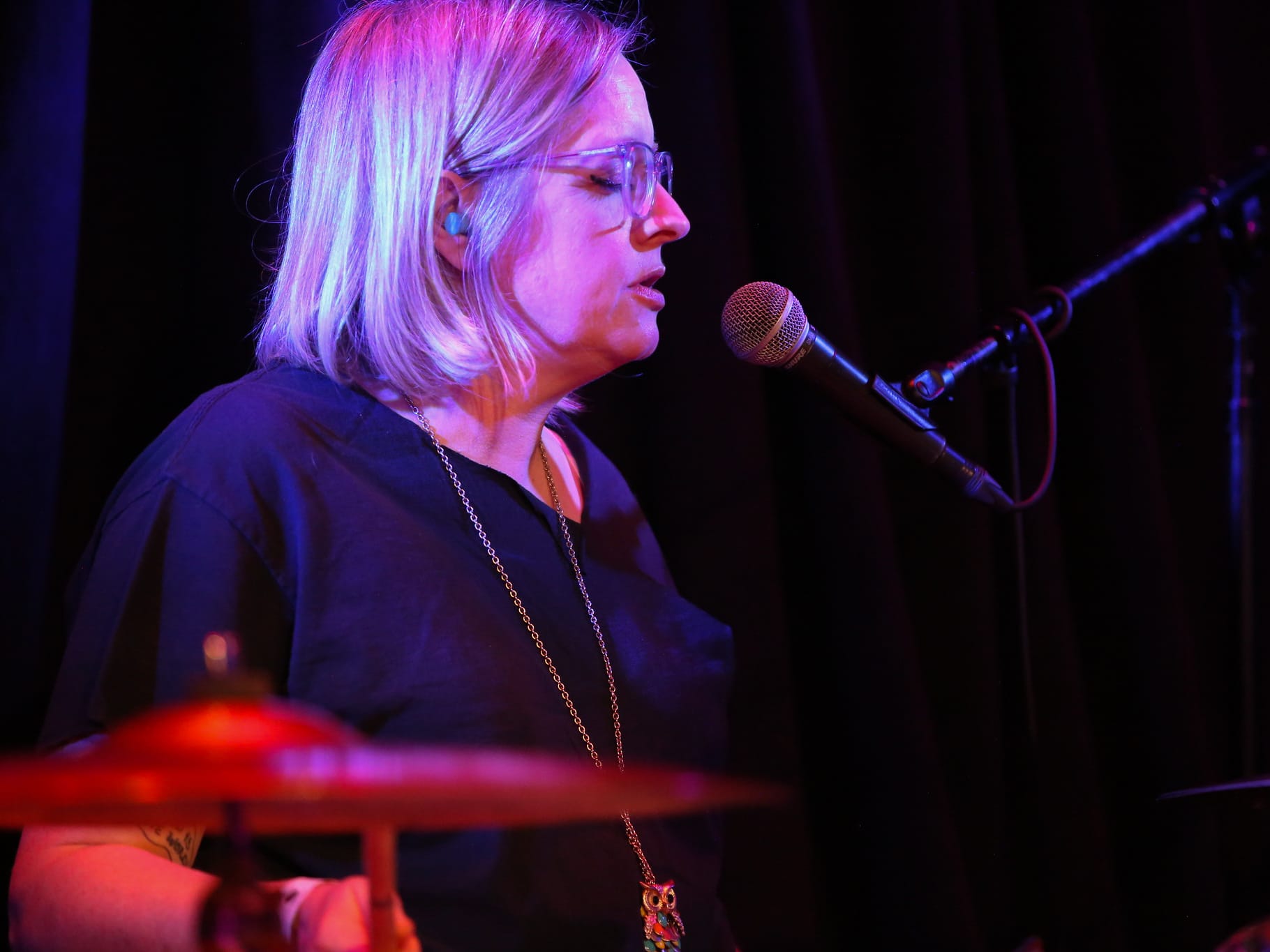 Drummer Heather Sawyer performs live at High Noon Saloon in 2024 during the farewell show of her former band, Proud Parents. She is pictured in profile. Her eyes are closed and she is singing into a microphone while drumming. She is pictured to the slight left of the image, a microphone extending in to take up the right. A ride cymbal is in soft focus in the lower left. Sawyer sports a blonde bob, glasses, blue earplugs, a dark v-neck shirt, and a gold chain that supports an owl.