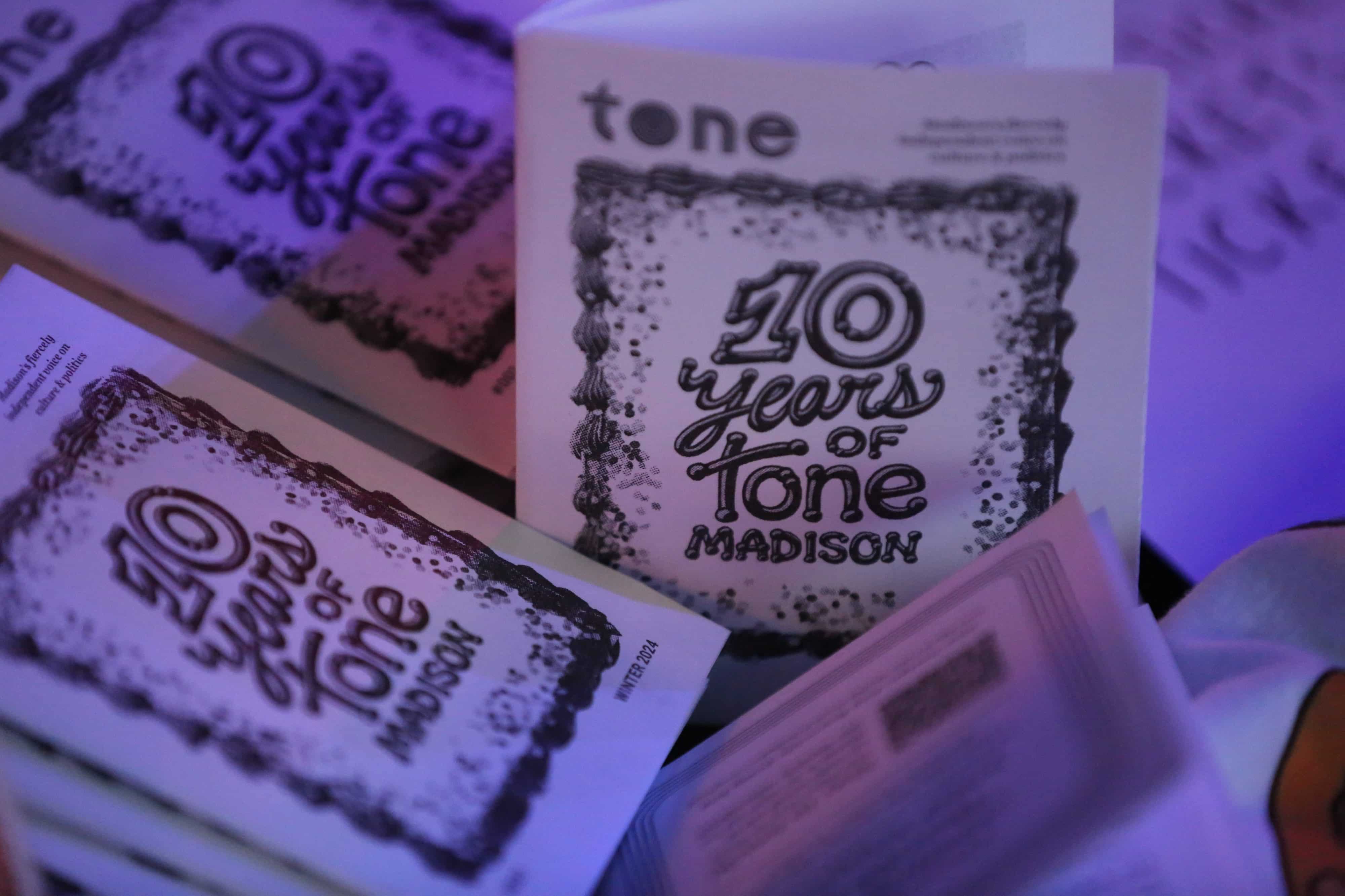 A table display of Tone Madison's first official print zine. A blue hue permeates through an image of a table full of print zines that show the Tone Madison logo and a poster-esque bordered illustration that reads "10 years of Tone Madison." The zine just right of center is propped and standing, in sharp focus, while a sea of the same zine surround it, just out of focus.