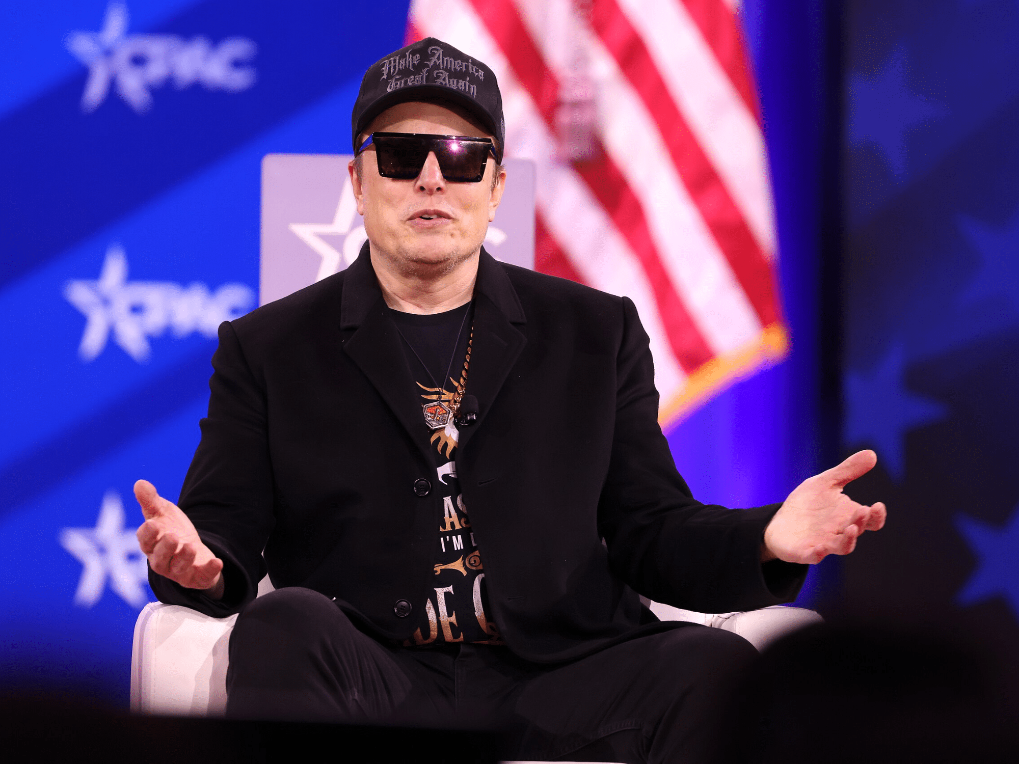 Elon Musk sits on a the Conservative Political Action Conference (CPAC) stage wearing a MAGA hat, square sunglasses and black blazer over a t-shirt.
