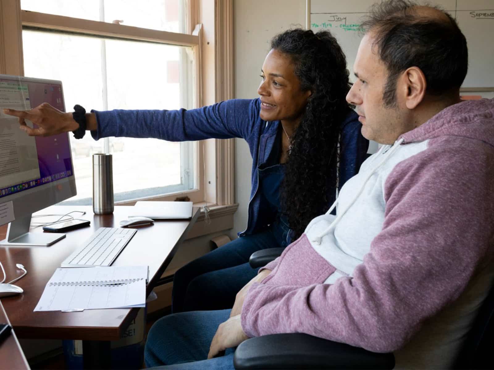 A photo shows a care worker sitting at a computer with a client. The care worker is pointing at the computer screen to show the client something.