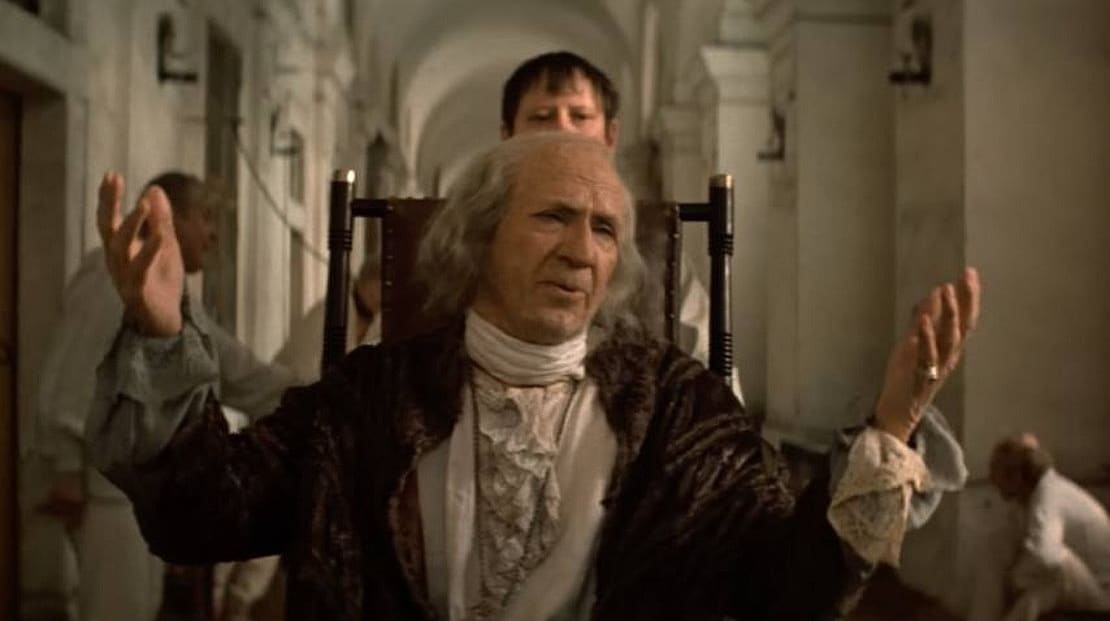 An aging man in slightly weathered 18th century clothing sits in a chair in an asylum hallway. He raises both his arms up in a pleading gesture and facial expression.