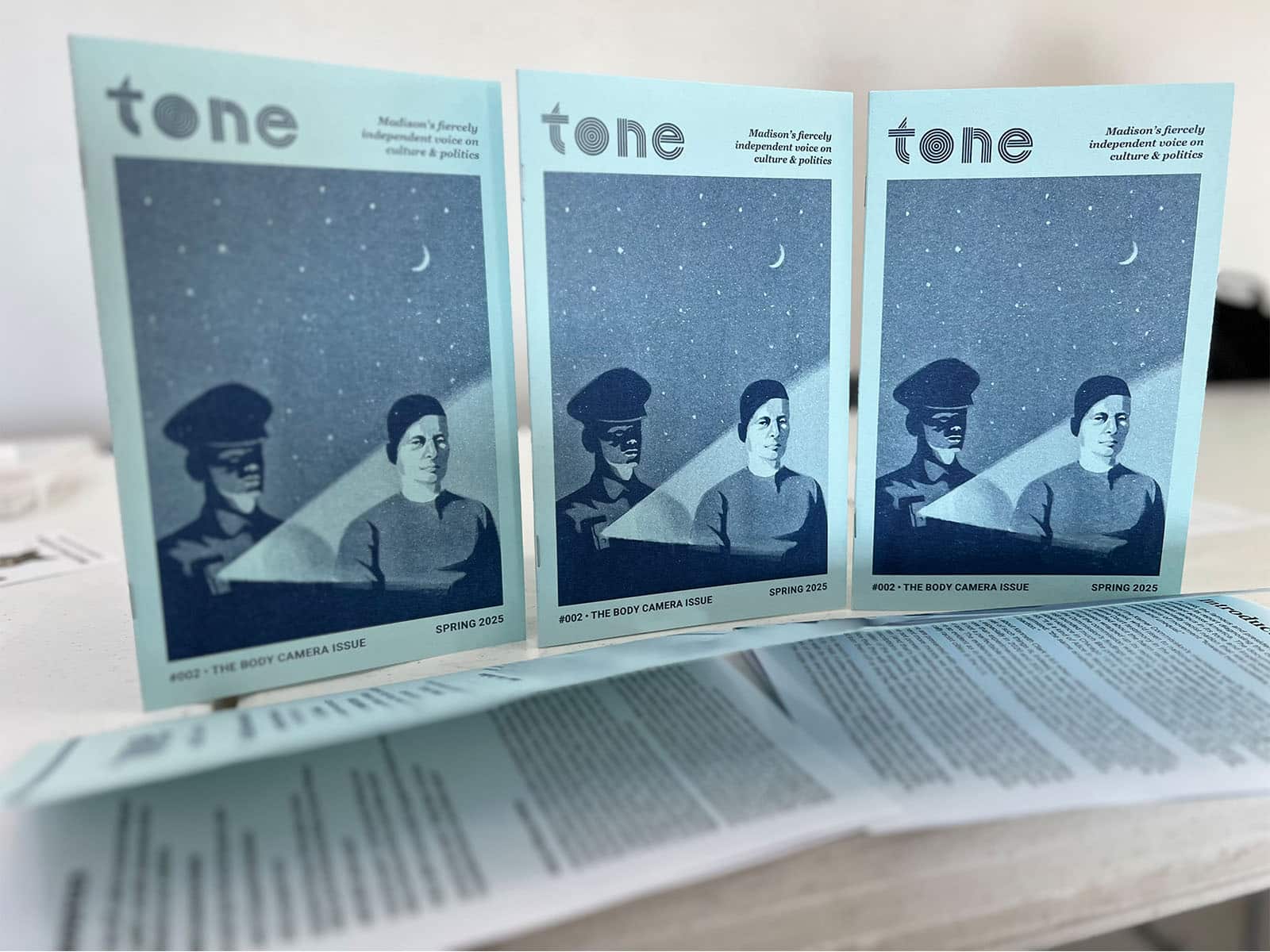 A photo shows copies of a printed small-format publication arranged in a row. They have blue covers printed with an illustration that depicts a police officer standing behind a man. A beam of light emanates from the officer's body-worn camera.