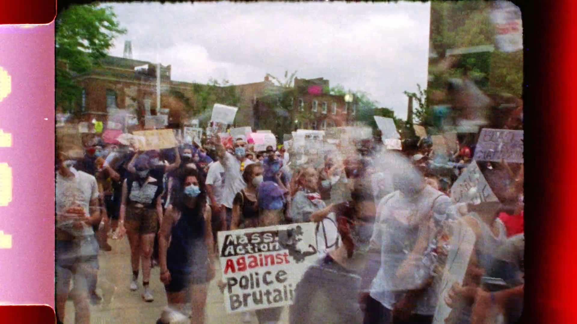 Slightly blurry and superimposed footage shows a crowd of masked protestors marching outside. Many of them are holding signs that speak to "Black Lives Matter" and against police brutality. The edges of the frame on the left and right sides have red/pink/black color flare aberrations from celluloid processing.
