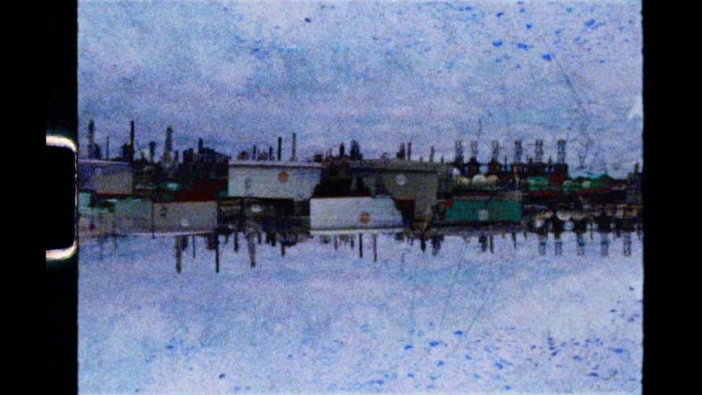 An image of an industrial cityscape is elongated at the center. The distant view of towers and stacks are mirrored and flipped on top of each other, producing a surreal effect. Above and below the cityscape is a hazy, distorted blue sky with blended shades of green and purple.