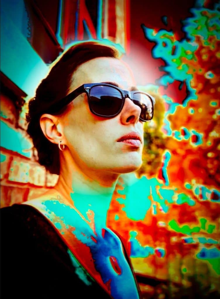 A photo with a high-contrast infrared-like effect shows a 1/4 headshot of filmmaker and artist Meggen Heuss. She stands outside near a building and trees behind her, distorted by the intensity of the red and green image effect. Heuss wears sunglasses, and stares upward at an angle.