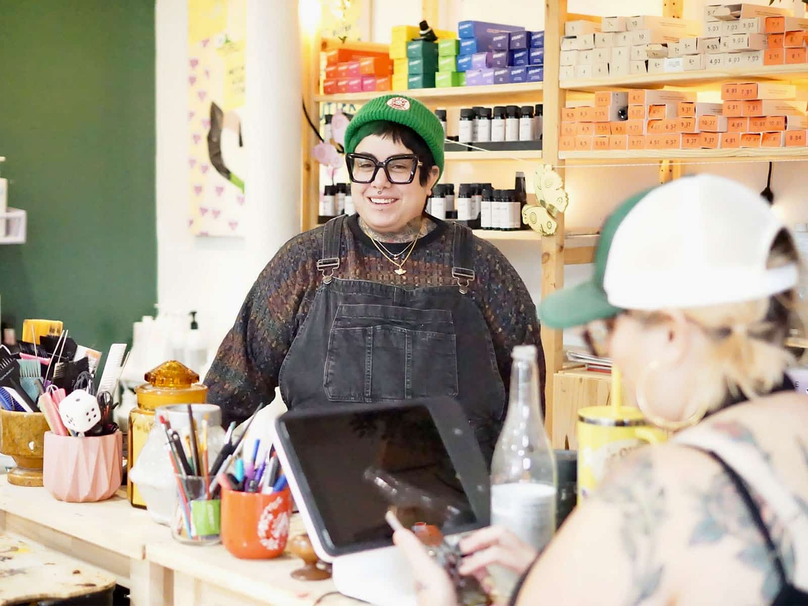 A photo shows El Ershler standing behind a counter at their hair salon, Mint Cuts and Color, talking with a customer who is visible out of focus. the foreground. The counter holds a tablet and cups full of pens and art supplies. Behind Ershler are shelves holding a colorful array of hair products.
