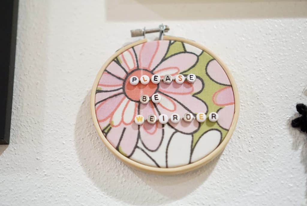 A photo shows a woven piece of art depicting a flower, decorated with letters that spell out "PLEASE BE WEIRDER."