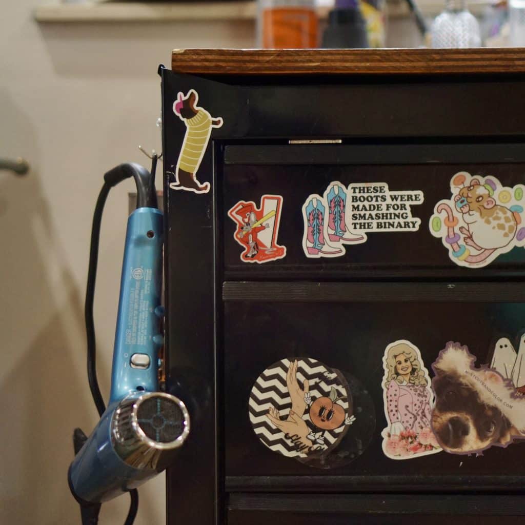 A photo shows a detail of a workstation at a hair salon. A toolbox-like piece of furniture is decorated with stickers, including one that reads "these boots were made for smashing the binary," and a hair dryer hangs from a hook on the side.