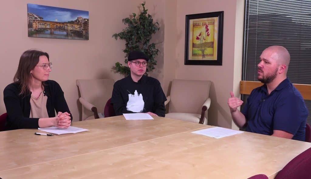 At a medium shot, three people sit in a conference room in a triangular-like formation at a large tan table. Two of them, sitting at the left and center, are turned towards the host, who sits at the right. They all have paper notes in front of them. The table consumes the entire foreground of the frame.
