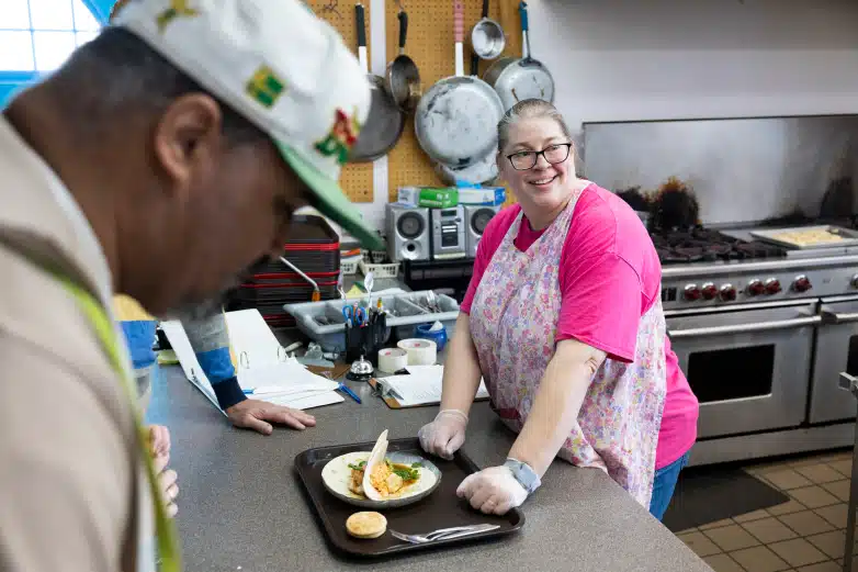 Woman wearing glasses and a pink shirt and apron smiles in a kitchen next to a tray of food on a counter. Two other people on the left