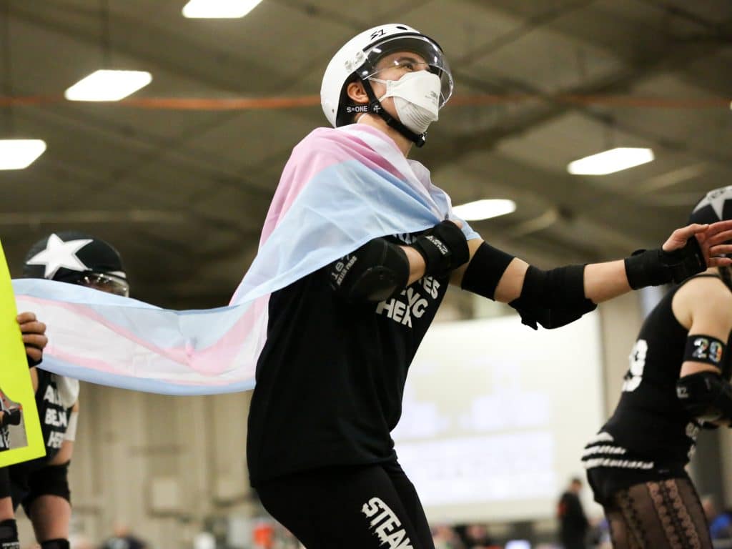 Emily Mills is shown roller skating during an intro rollout for the Madison Roller Derby home team the Reservoir Dolls. They are wearing a mask, a white helmet, and an all-black uniform. Their shirt, though partially obscured in the photo, reads "Trans Athletes Belong Here." They seem to be smiling behind their mask. They have fashioned the trans flag as a cape, and it's swept up in the wind behind them. One of their hands is clutching the flag's ends together as they skate, while the other is outstretched. They're wearing a white helmet and are in mid-stride, surrounded by a few of their Reservoir Doll teammates.