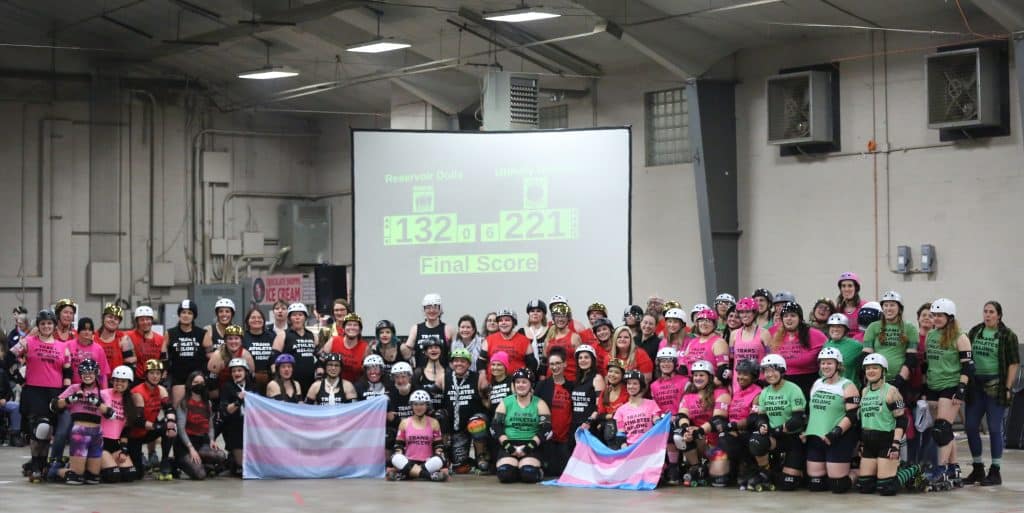 Madison Roller Derby's four home teams pose for a group photo. Nearly every member of the league is wearing a shirt that reads "TRANS ATHLETES BELONG HERE" in bold black letters. There are around 80 league members in the photo. A few of the skaters in the front rows are holding up trans flags and smiling.