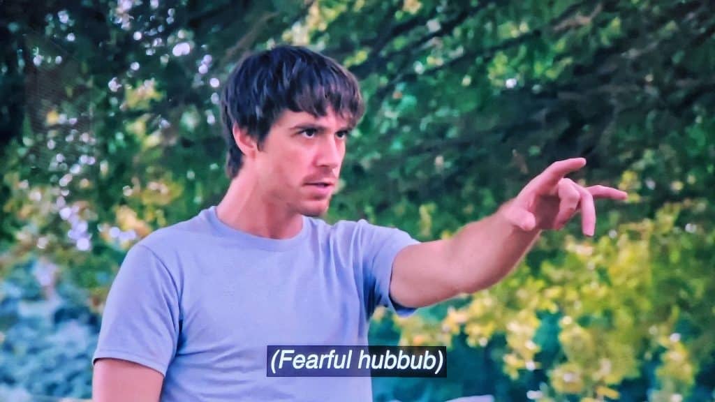 An impressionistic-looking photo of a television screen features a male actor standing outside staring off-screen to the right with an ill-natured facial expression. His hand is raised as if he's pointing or taunting. A subtitle on the screen near the bottom of the screen reads "(Fearful hubbub)."