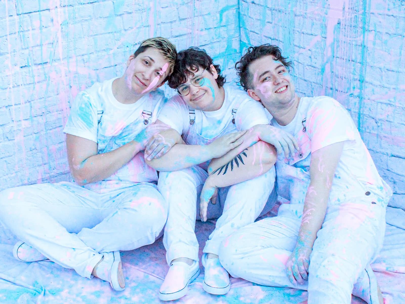 The three members of Kat and the Hurricane are seated in a once-white room, with all three members wearing once-white clothes. Both the walls and the members themselves have been splattered with pastel blue and pink paint. The band's members have their arms hooked around each other and are smiling at the camera.