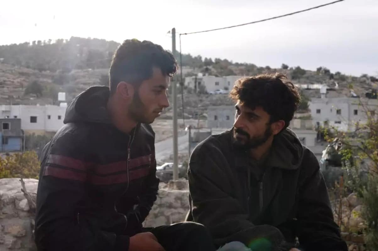 A still from the political documentary "No Other Land" shows two men sitting outside in the foreground. They wear jackets and turn towards one another with tentative expressions. White structures behind them extend into the hills in the distance along with trees and a bright white sky.