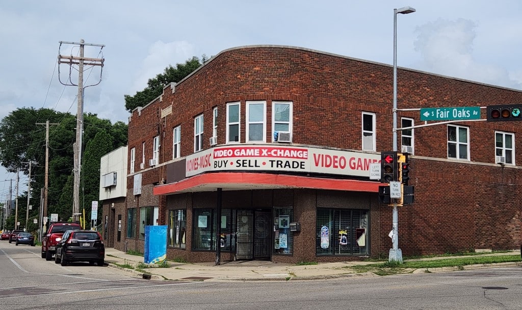 A photo shows the building and storefront of Video Game X-Change at the corner intersection of S Fair Oaks Av and Atwood Av in Madison on a summer afternoon. The large store sign wraps around the side of the building, reading 