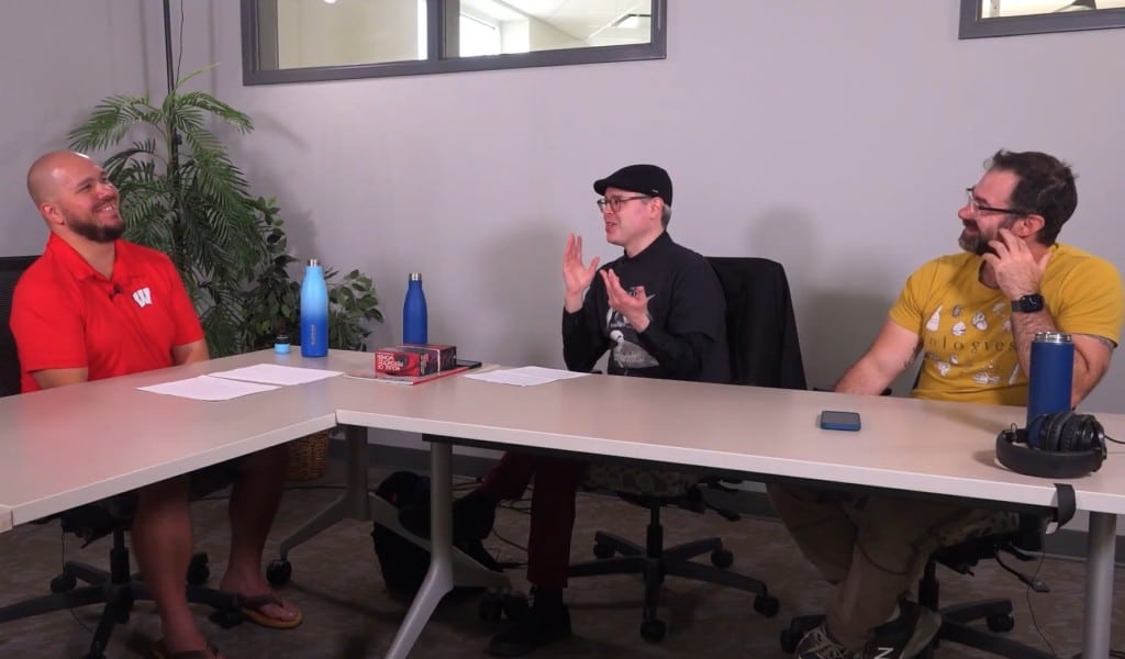 At a medium shot, three people sit in a simple conference room at two elongated desks arranged in a triangular-like formation. The two people on the right are turned towards the host on the left, smiling jovially. Paper notes, steel water bottles, and a phone are placed on the tables in front of or to the side of the three people.
