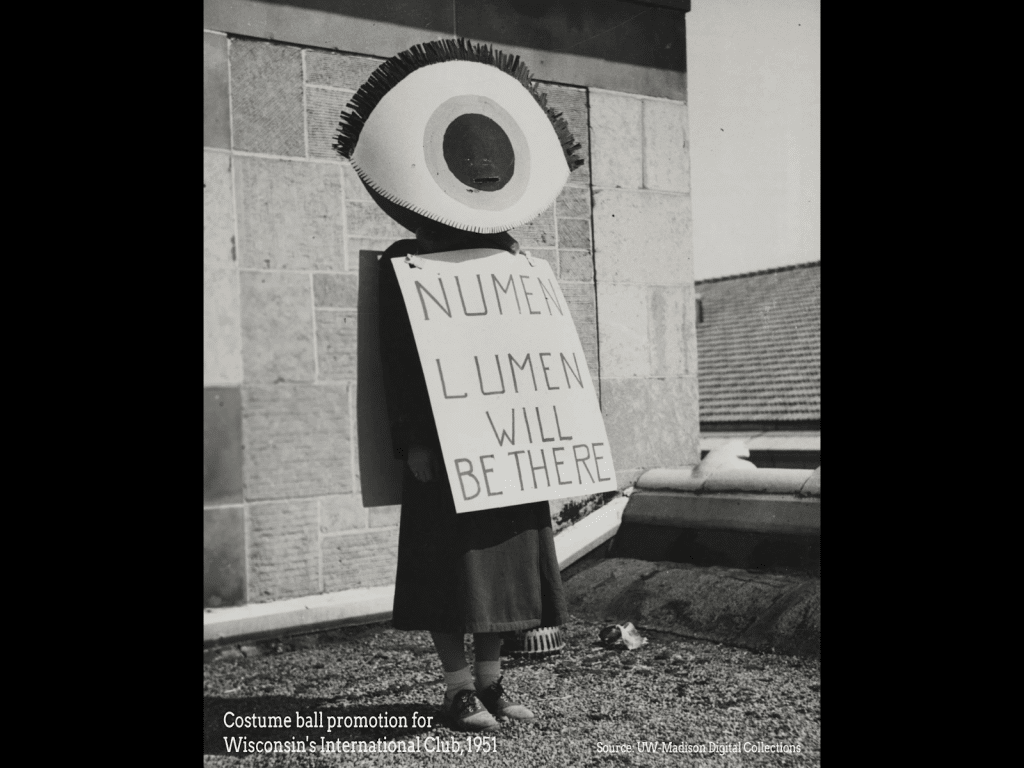 A historic black-and-white photograph shows a female body wearing a giant eye that covers their head and a posterboard sign that reads 