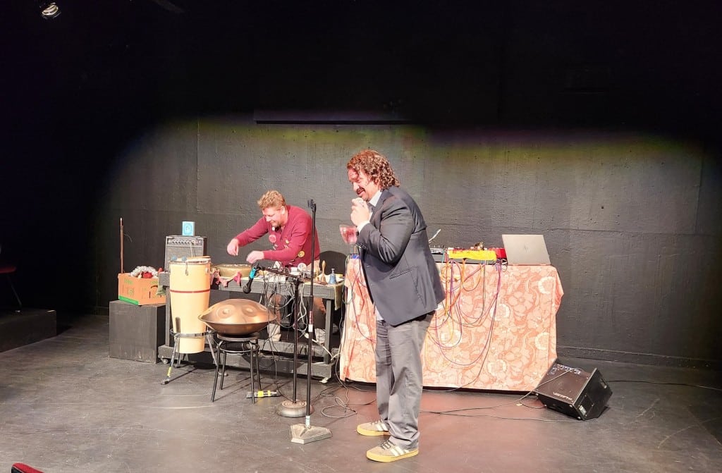 Photo of a black stage with two performers standing in front of and behind tables with a complex, amplified audio setup. On the right, a man wears a suit jacket and holds a microphone in his left hand while leaning in to speak. Behind him to the left, another man wears a red sweater with numerous buttons pinned to it. He is gently tossing small objects onto a flat, gold-colored surface.