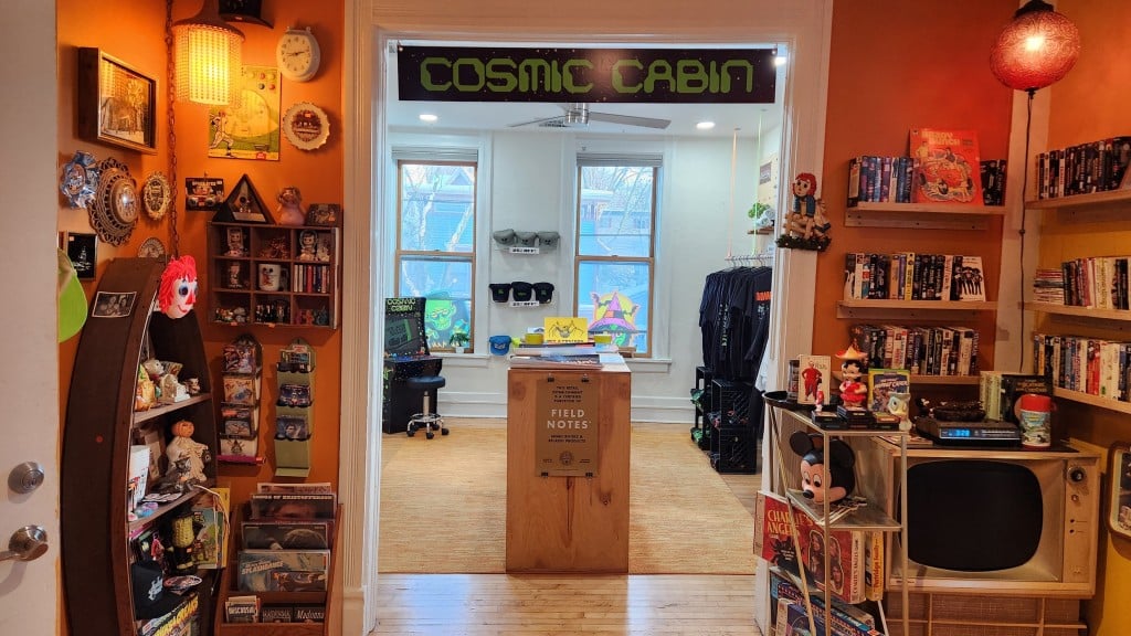 A photo shows various collectible paraphernalia—toys, records, VHS tapes, board games, etc.—along the walls in a mood-lit foyer. In the center is a wide entrance that opens into a brighter sunlit room that extends into the background. The back room displays racks of t-shirts and some hats along the walls. Posters are arranged flat on a table in the middle of the back room. A sign hangs above the entrance, reading 