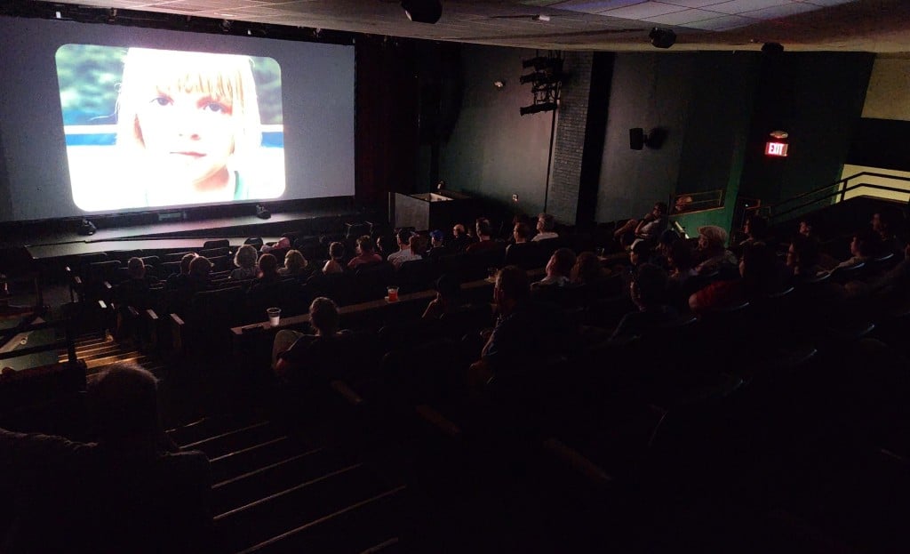 Photo of a darkened movie theater at an angle captures a crowd of people watching the illuminated screen at the top left, which shows a close-up of the face of Arabella Olivia Clark as Addie in the short film 