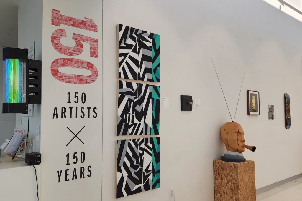 A slightly angled photo of an art gallery space with white walls. Various artworks large and small are displayed on the wall (and one a wooden pedestal on the marble floor). At the left, the number 150 is displayed vertically in a large pinkish-red font, while the exhibition name