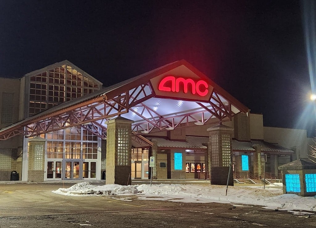 A slightly angled photo shows the entrance to an AMC movie theater at night. The large metal awning extends out a couple dozen feet from the main doors with the AMC logo illuminated in red.