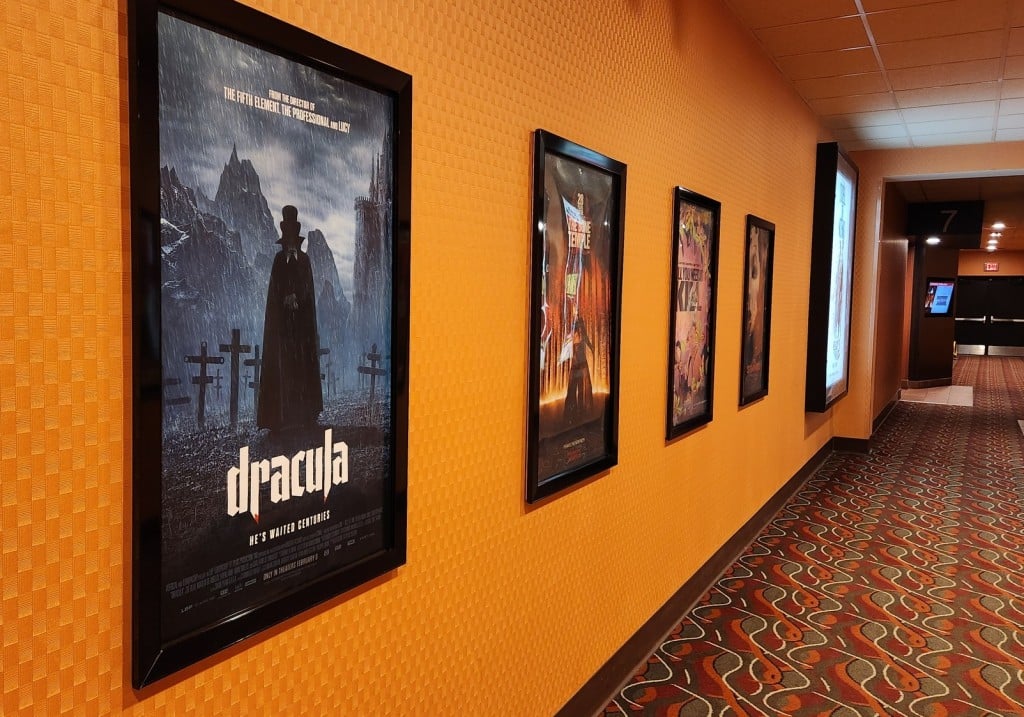 An angled photo in a movie theater hallway displays a row of framed posters hanging on the orange wall. The posters include