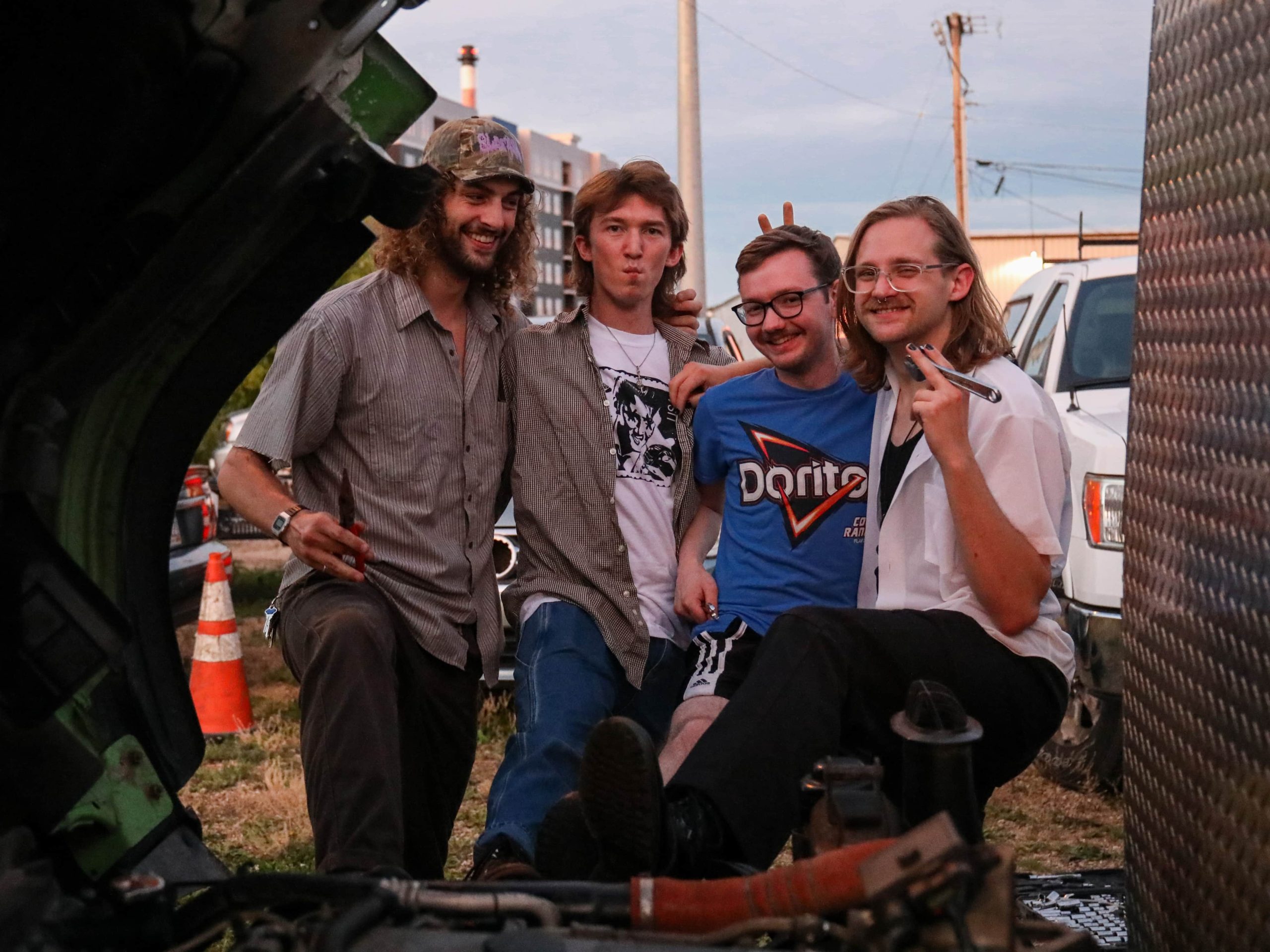 Slick pose with their arms around each other in front of a vehicle being serviced at an auto repair shop. From Left to Right: Calvin Childress, Nate Opperman, Hance Throckmorton, and Morgen Nicodemus. Each band member is holding a small repair tool. They're all smiling, apart from Opperman, whose lips are pursed.||