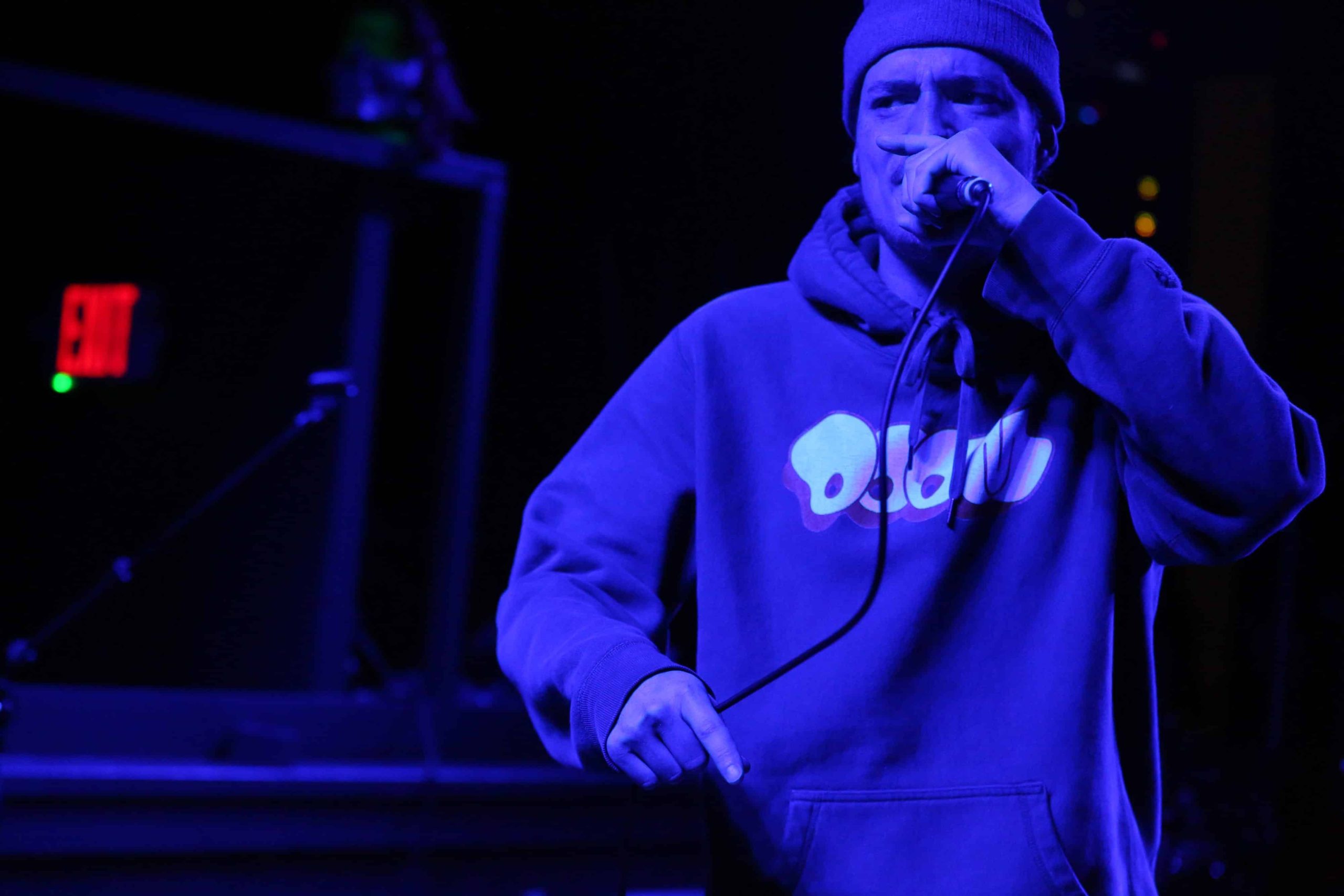 Alex Driver performs at Gamma Ray Bar as Fred Really. He's wearing a beanie and an MF DOOM hoodie. He's shown in deep concentration, gripping the microphone while rapping. Driver takes up the right half of the image, bathed in dark blue stage lights.