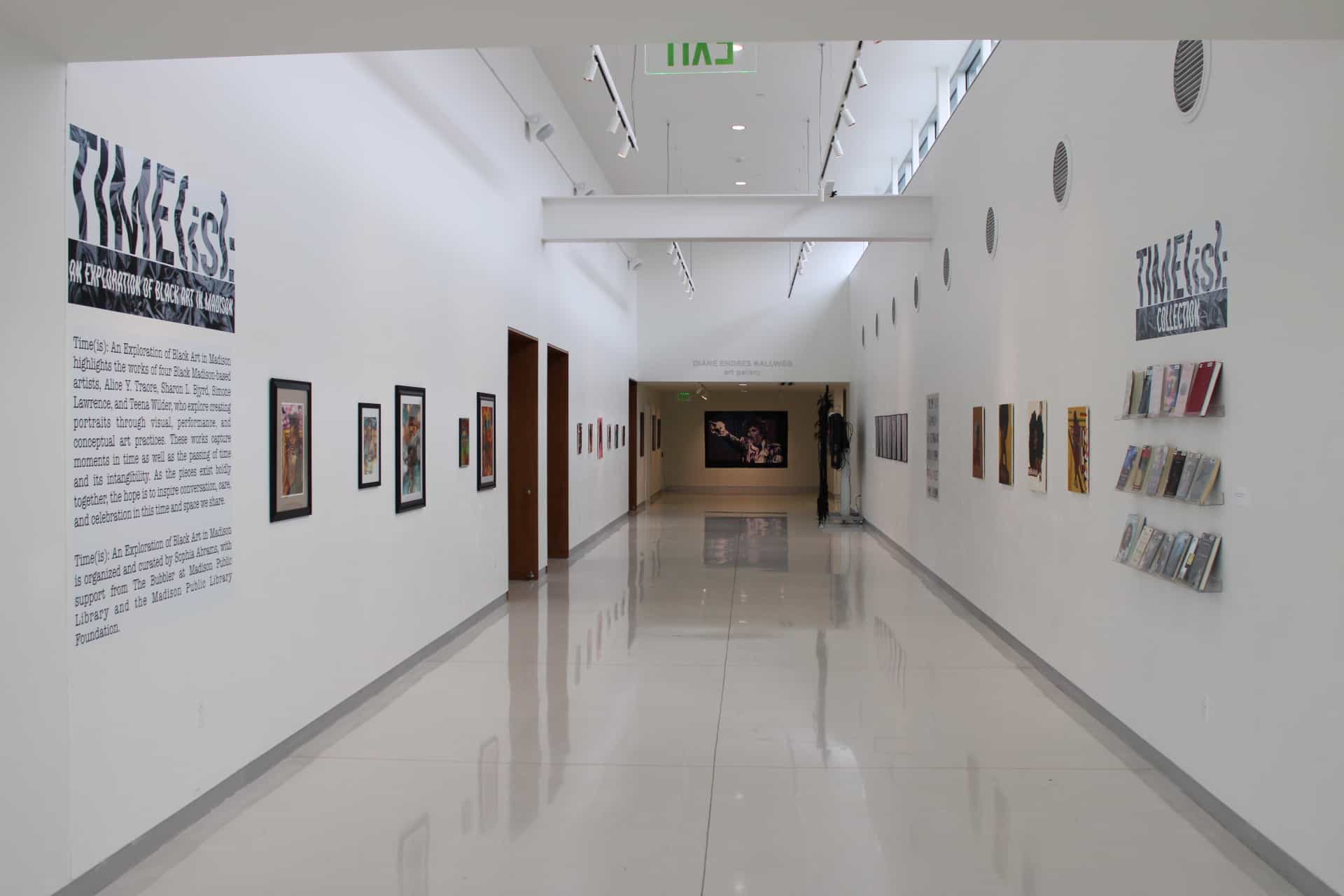 A view of the "Time(is)" show in the gallery on the third floor of the Central Library in downtown Madison. The gallery is a long hallway; in the foreground, to the left, is a block of text printed on a wall introducing the exhibition. On the right are shelves of books associated with the exhibition. Prints and paintings stretch down the length of the gallery on either side.||