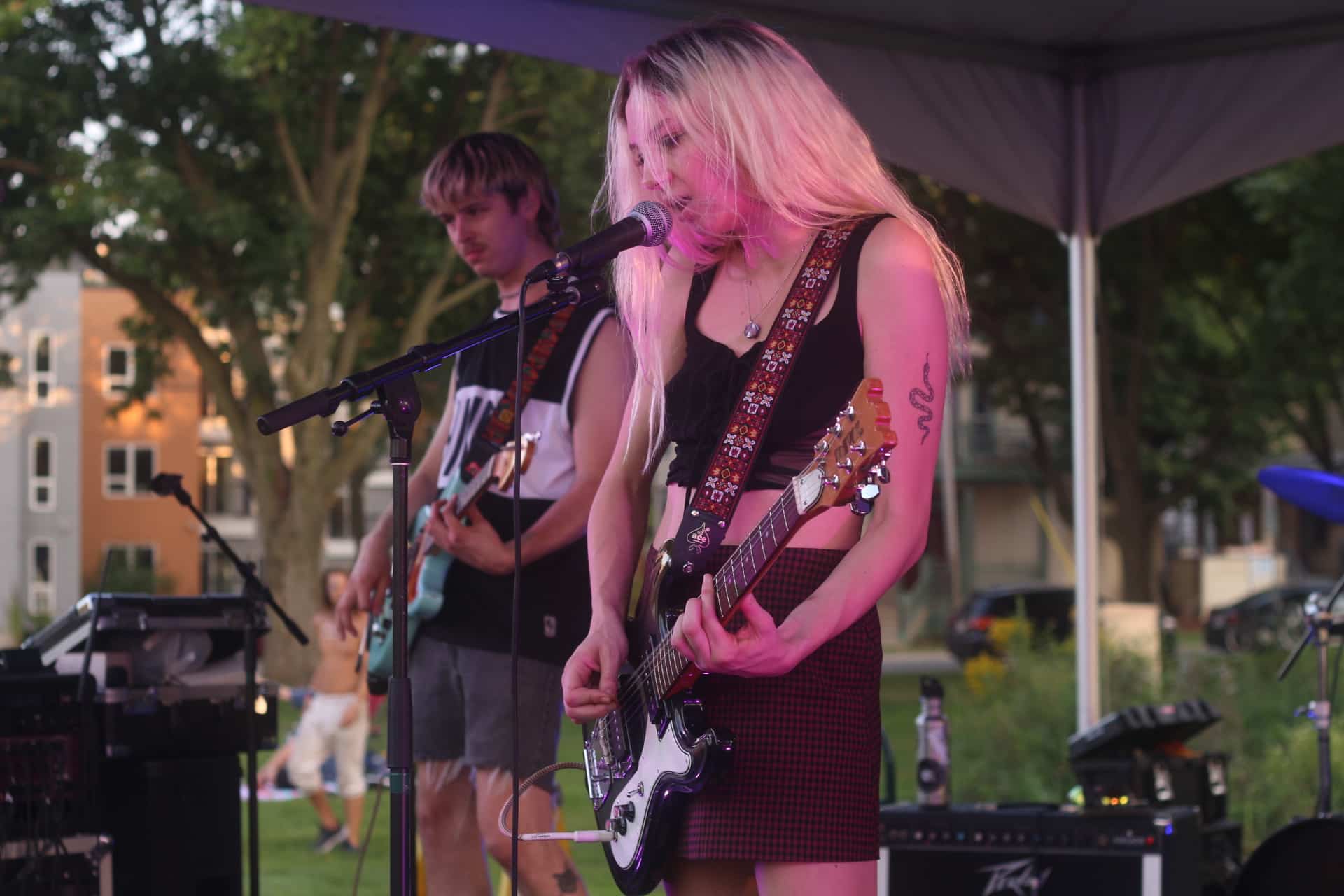 Interlay performs as part of WSUM's Party In The Park 2022 event. Guitarist/vocalist Alexandria Ortgiesen is centered, singing through wisps of windswept hair, guitar slung low. Guitarist Alex Edlund is in soft focus behind her, to the left. Buildings and trees can be seen in the background.