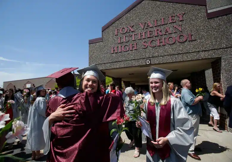Graduating students in caps and gowns hug in front of the entrance to Fox Valley Lutheran High School.