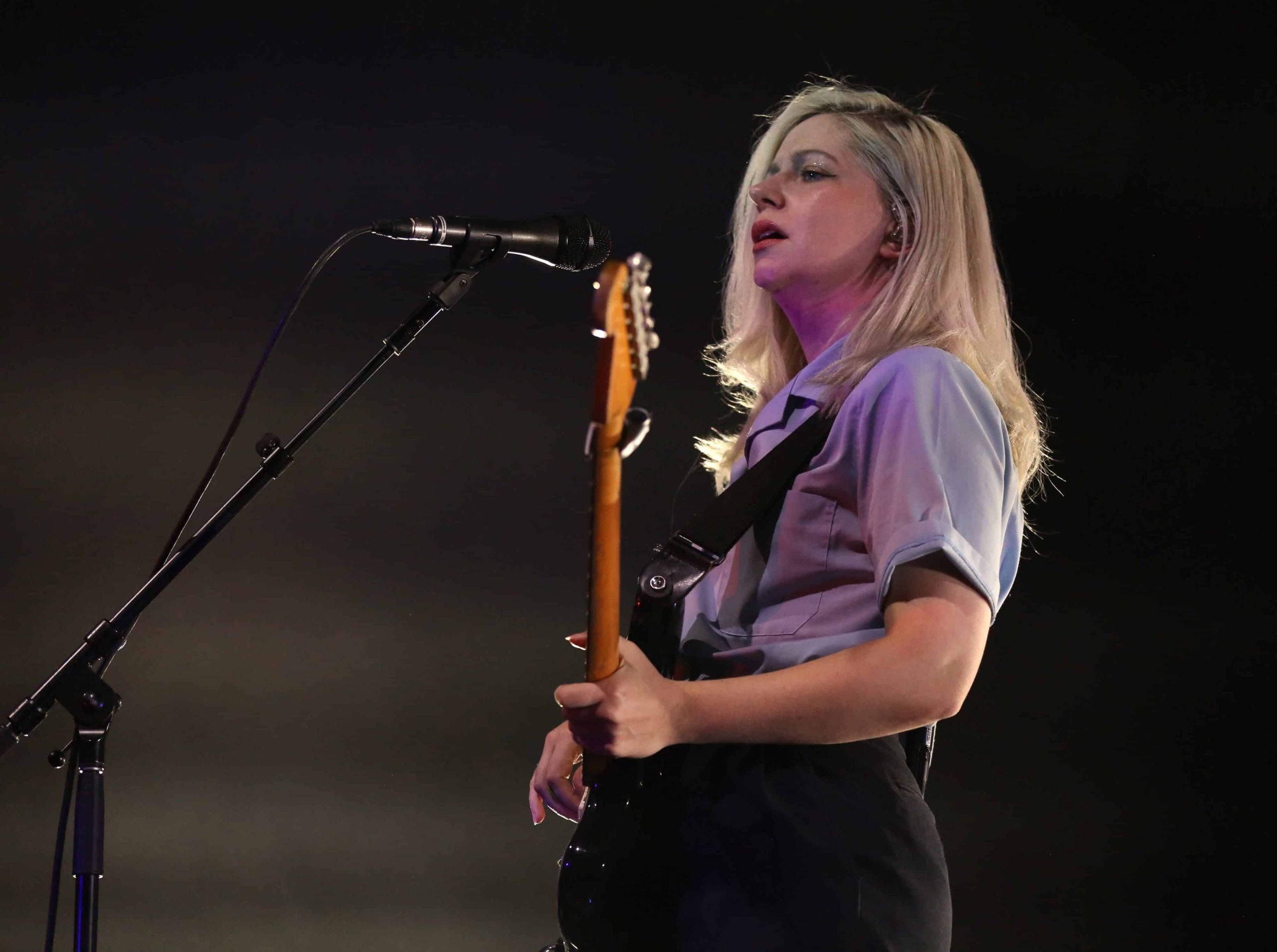 Alvvays guitarist/vocalist Molly Rankin approaches a microphone while looking out over the audience. She's wearing a baby blue button-up t-shirt and black pants. She is in profile, positioned to the slight right of the image, while the microphone is slightly left. The background behind her is dark, but she is back-lit by stage lighting, creating a soft glow effect.|