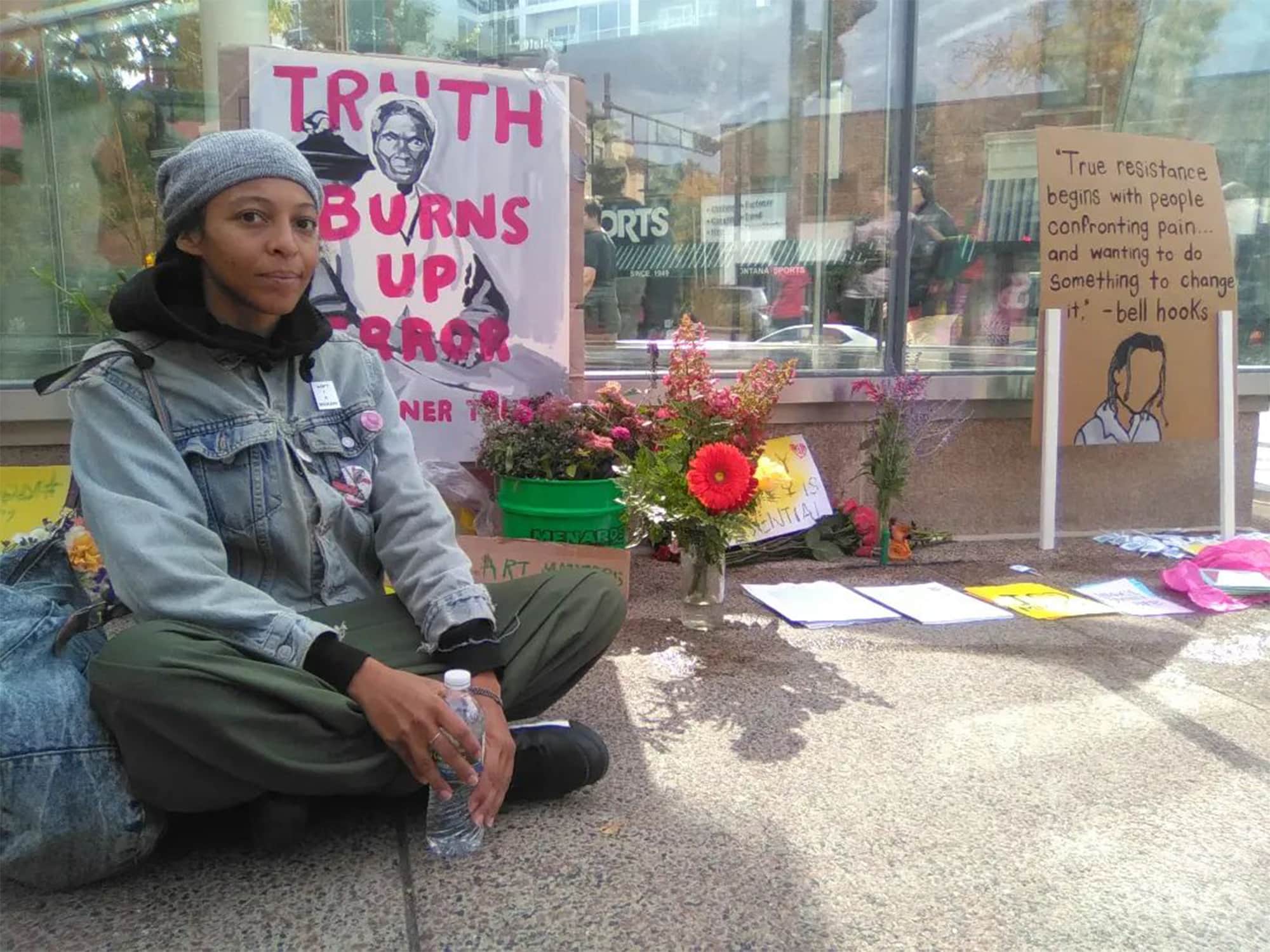 A photo shows artist Mars Patterson sitting on the sidewalk in front of the Madison Museum of Contemporary Art. Behind them are signs, flowers, and other materials displayed as part of a protest. One sign reads "Truth burns up error—Sojourner Truth." Another reads "True resistance begins with people confronting pain... and wanting to do something to change it. —bell hooks." The glass exterior of the museum lobby is also visible in the background.|||