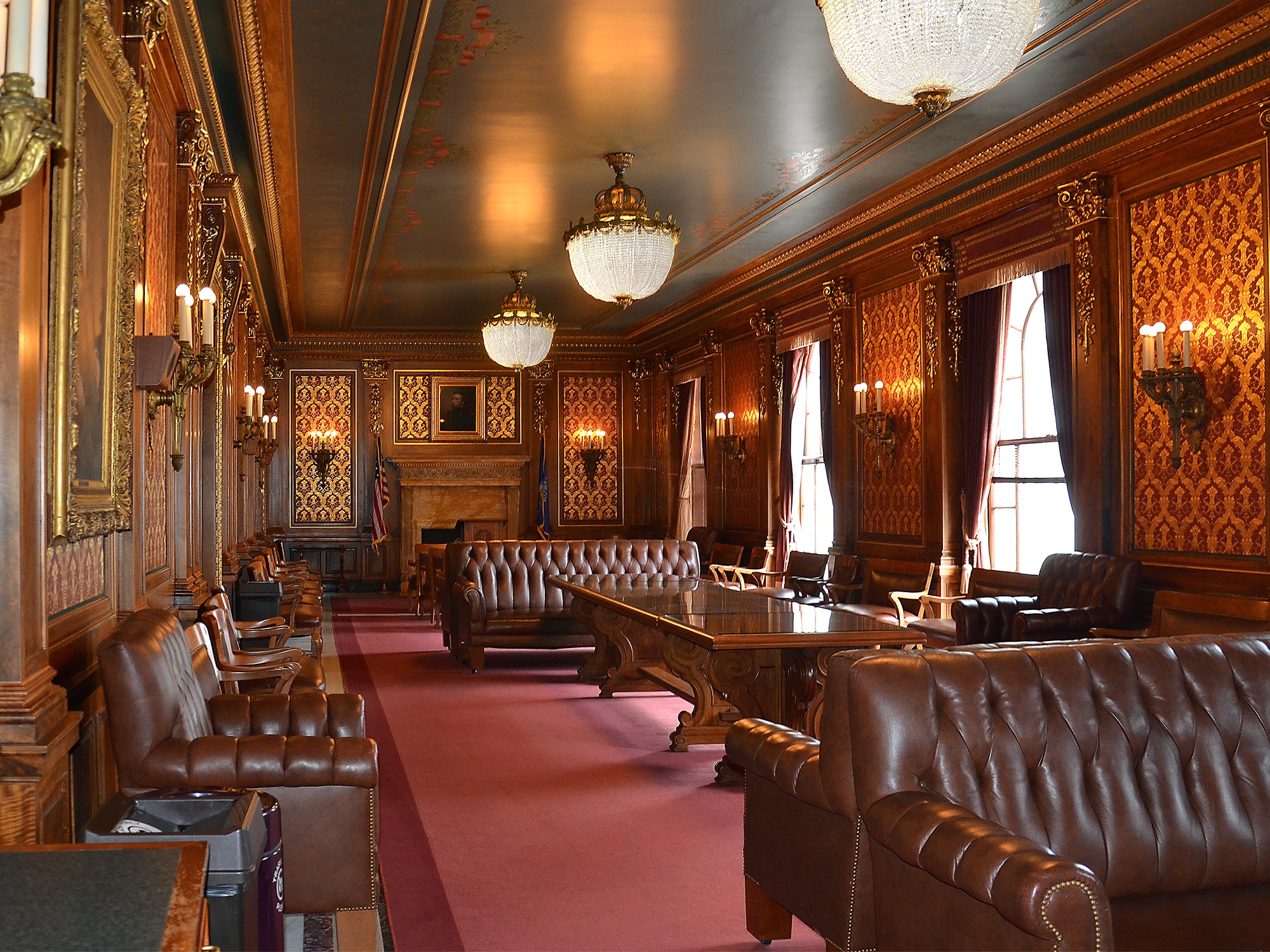 A photo shows a parlor in the Wisconsin State Capitol. The room has red carpet and a number of upholstered, expensive-looking couches, with ornate lighting and decorations on the walls.