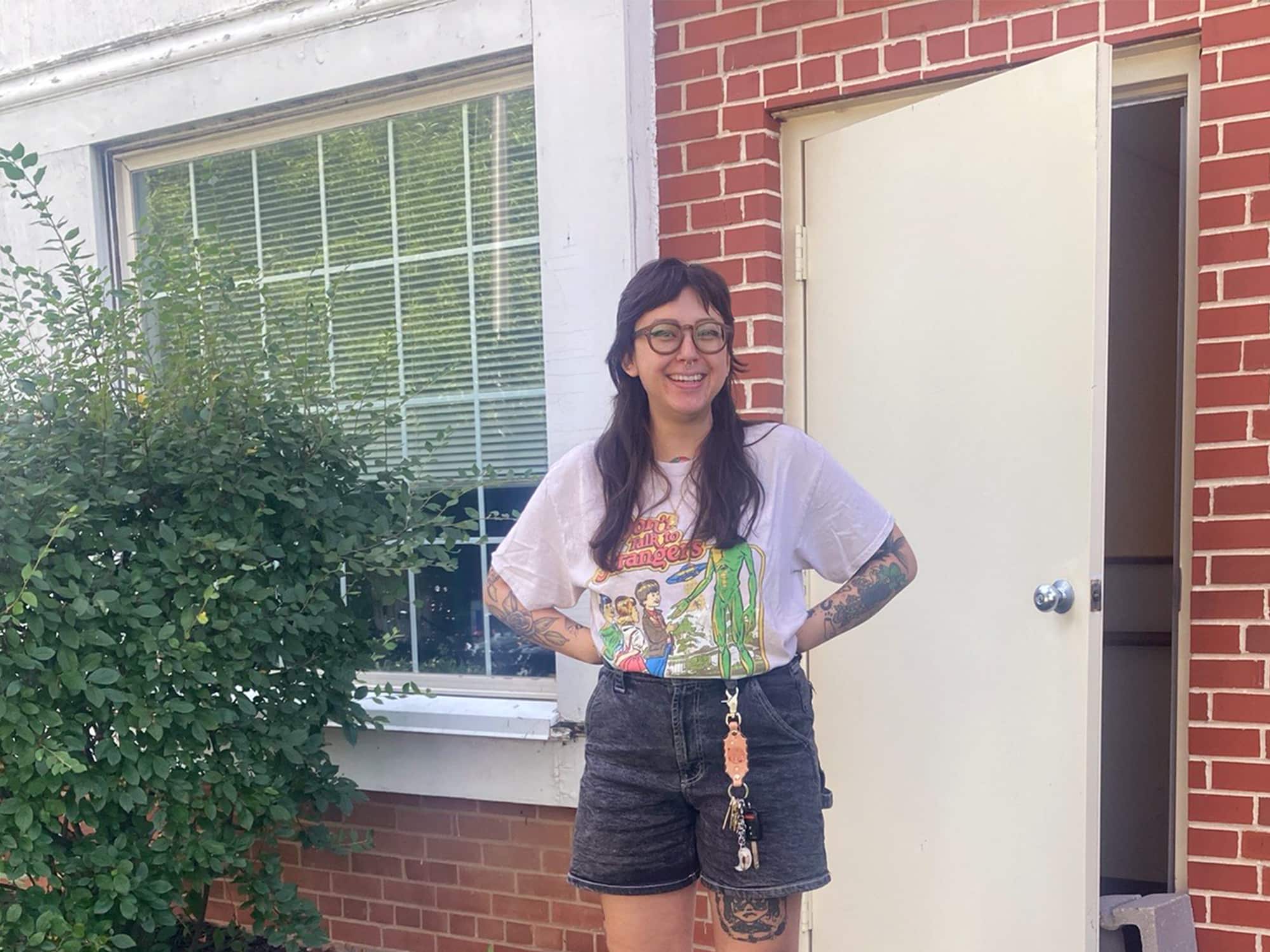 Maggie Denman is shown standing in the doorway to the office building where her shop Boneset Records will operate. Behind her is a brick wall with a partially opened door in it, and to her left is a window and a large shrub.
