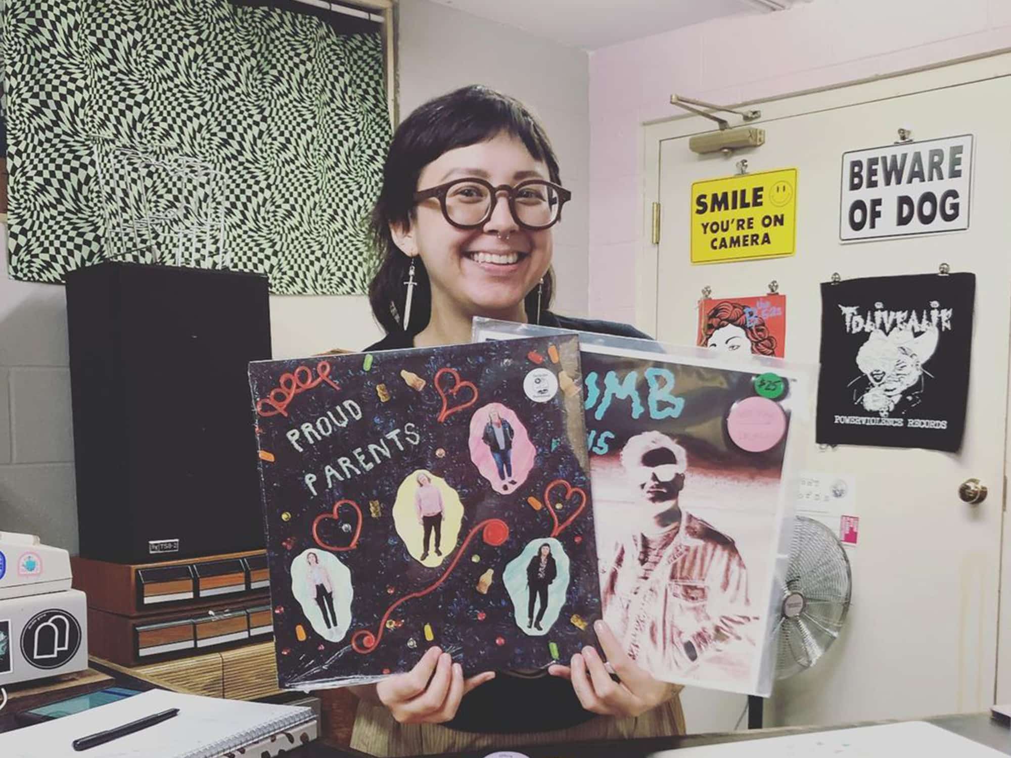 A photo shows Boneset Records owner Maggie Denman standing behind a counter and holding up two LPs, one from Proud Parents and the other from Baby Tyler. Behind her is a speaker, an organizer for cassette tapes, and wall signs with messages that include "smile, you're on camera" and "beware of dog."