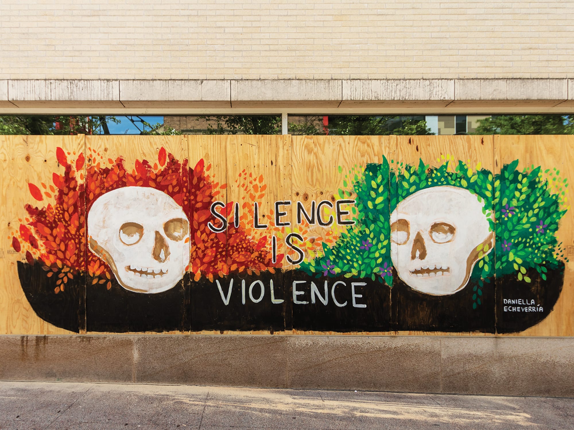 A photo shows Daniella Echeverría's mural "Silence Is Violence," painted on plywood boards covering broken windows at the Madison Museum of Contemporary Art. Above the boards, a few inches of the windows are still visible, and above that part of the white brick facade of the building. The mural consists of two skulls, one surrounded by green plants and one surrounded by red and orange plants. In between the two skulls, the words "SILENCE IS VIOLENCE" are written in large capital letters.