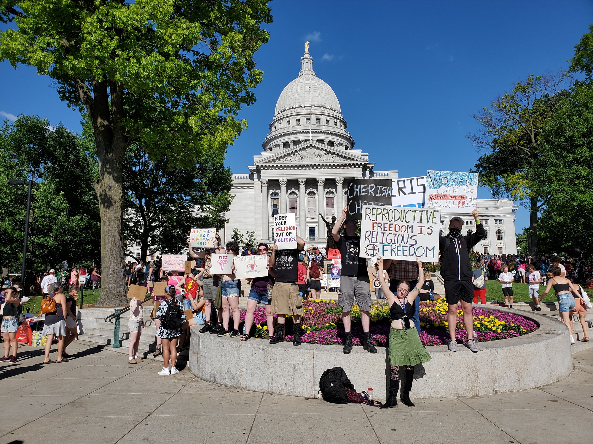 Demonstrators at a Friday, June 24 protest at the Wisconsin State Capitol stand on a circular planter on the Capitol grounds, holding up handmade signs with messages including “Reproductive freedom is religious freedom” and “keep your religion out of your politics.” photo by Emily Mills.