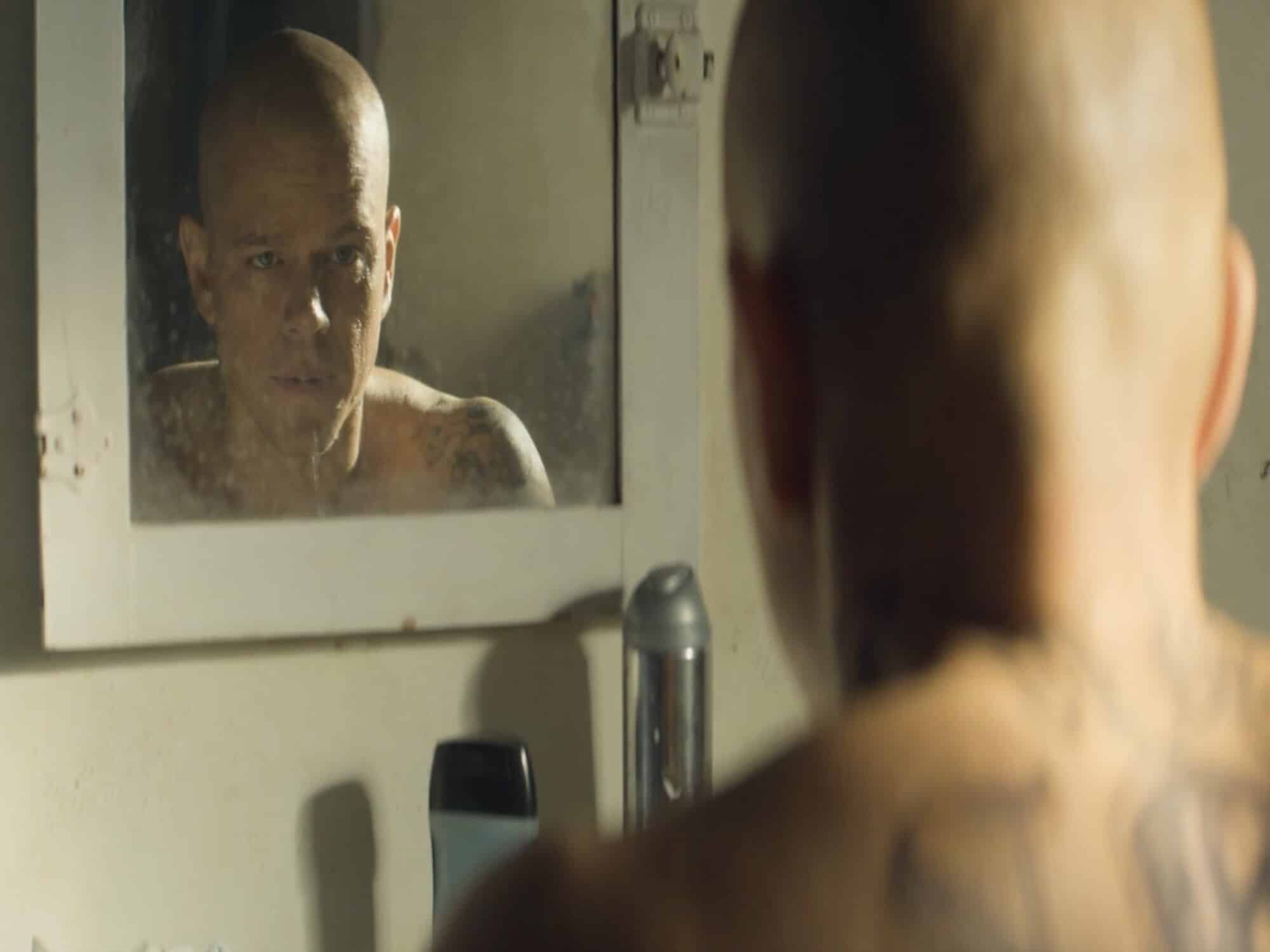 An over-the-shoulder screenshot from "Elysium" shows a bald thirtysomething man (Matt Damon) with a back tattoo staring into a smudgy bathroom mirror.