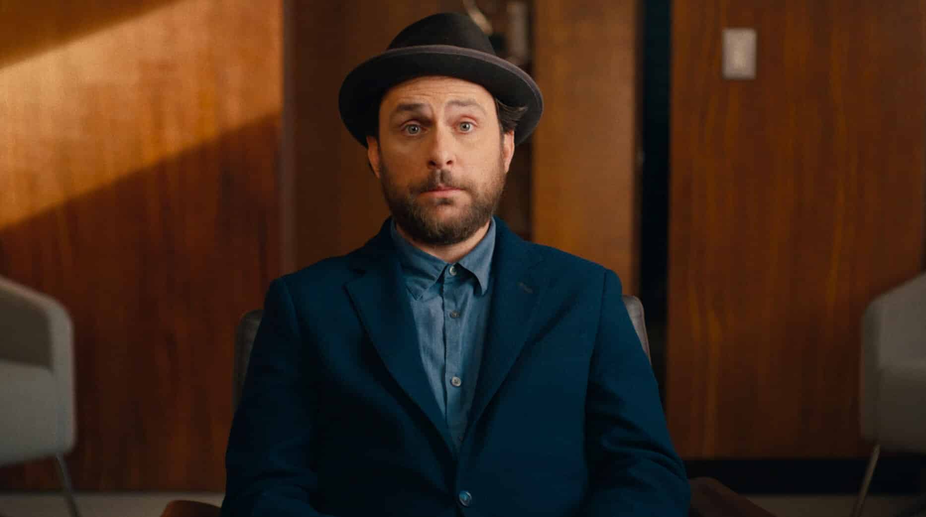 Wearing a blazer and fedora, Latte Pronto (Charlie Day) sits in an office and stares forward with a confused look on his face.