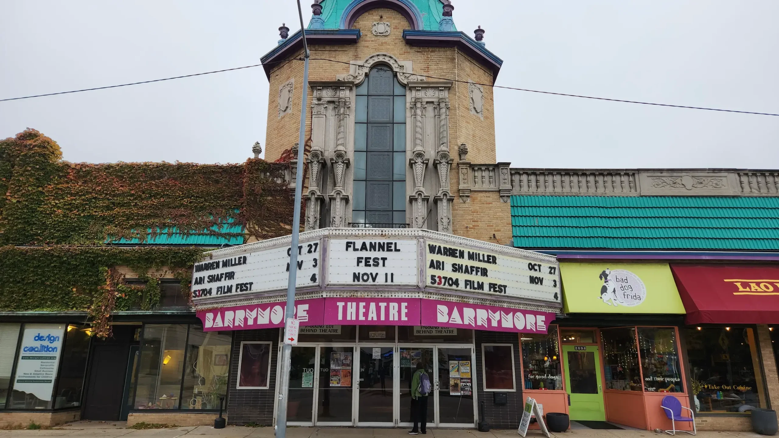 A slightly wide-angled, overcast photograph of the 2000 block of Atwood Avenue across the street from the Barrymore Theatre, which is centered in the frame. The black lettering on the marquee below the vertically ornate stone building displays several event dates with the 53704 Frame By Frame Film Festival at the bottom on Saturday, November 4.