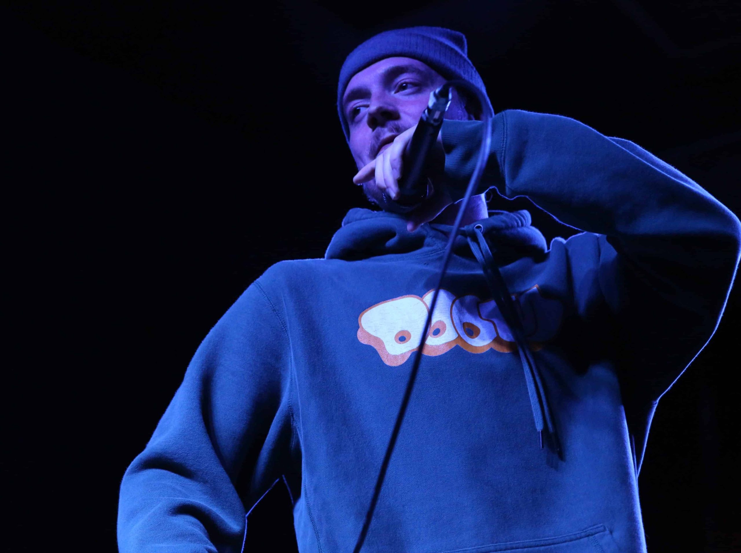 Rapper Fred Really performs during Tone Madison's 10th anniversary party at Gamma Ray Bar. He's cast in a cool blue stage light, wearing a DOOM hoodie and beanie. The microphone is near his face. He's positioned to the slight right and takes up the majority of the image. The backdrop is pitch black.