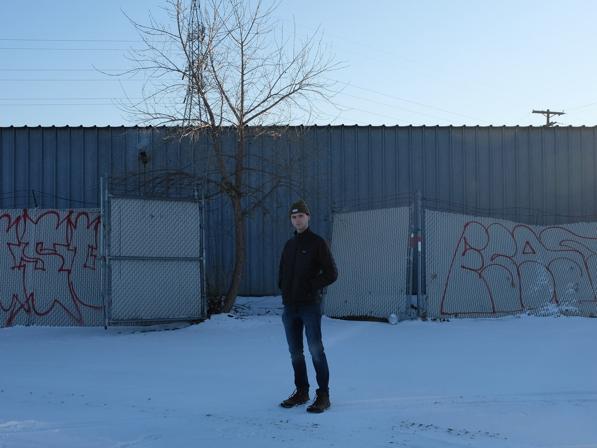 A publicity photo shows musician Hendrix Gullixson standing in the snow, in front of a fence and a metal building.