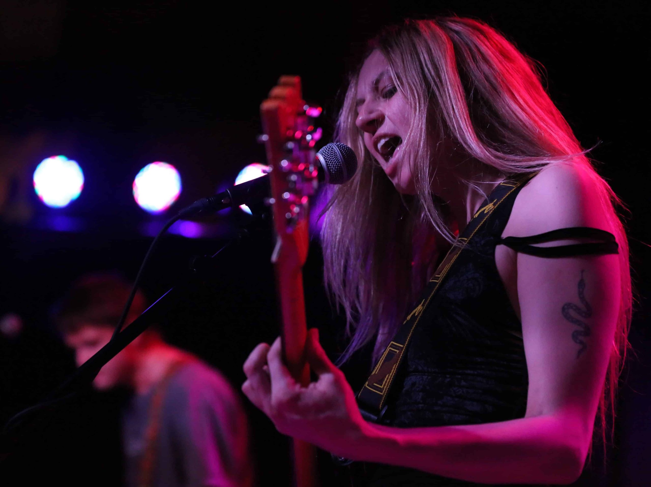 Interlay's Alexandria Ortgiesen snarls into a microphone while playing guitar at the High Noon Saloon. She takes up the right half of the image. In soft focus, distantly behind her to the left of the image is guitarist Sam Eklund. A small set of round stage lights are visible in the upper left.