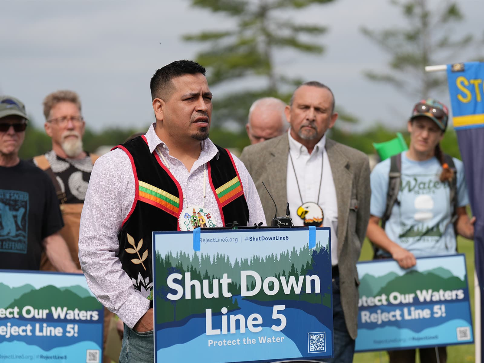 Daniel Wiggins Jr. deputy director of the Bad River Tribe’s Department of Natural Resources and others hold signs reading "Shut Down Line 5" as they speak at a press conference.|Screenshot of a map of of the current Line Five route in purple and the proposed reroute in red.|Rene Ann Goodrich, a Bad River Band Tribal Elder, speaks at a press conference.||Photo of a large room with two podiums at the from with one person speaking. The audience is seated on three other sides of the room.|Photo of a large room with two podiums at the from with one person speaking. The audience is seated on three other sides of the room.