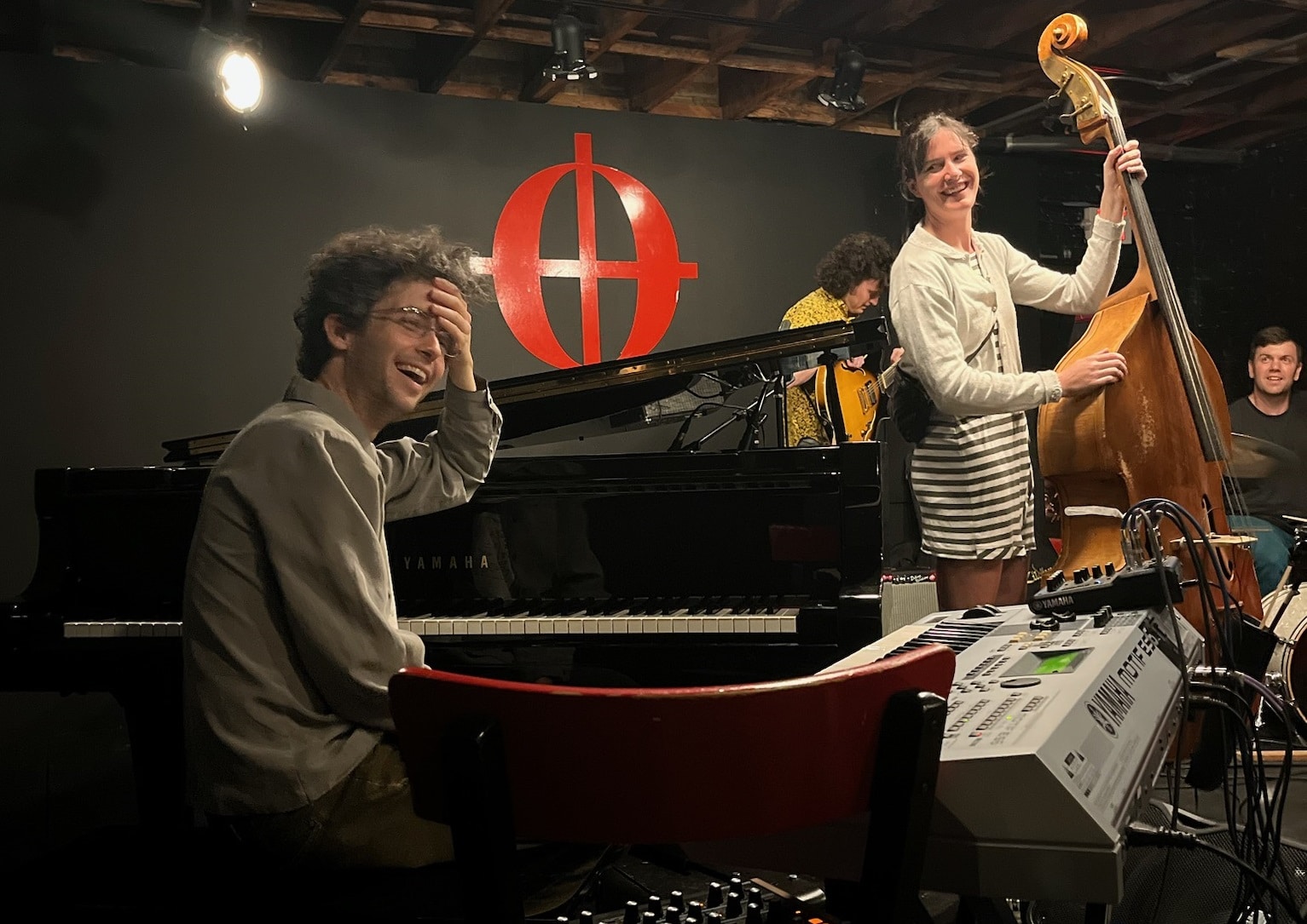 Four musicians on the Café Coda stage with black walls. Luke Leavitt sits at the grand piano to the left of center, laughing joyfully, with his left hand to his forehead. Bassist Claire Kannapell, electric guitarist Ryan Flannery, and drummer Tim Russell stand or sit behind him to the right, all smiling.