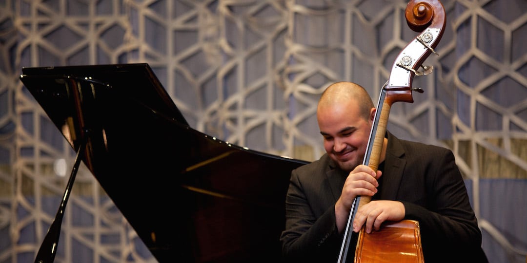 A photo shows musician Nick Moran standing with an upright bass and smiling, with his face angled a bit downward. Behind him is the open lid of a grand piano and a curtain.