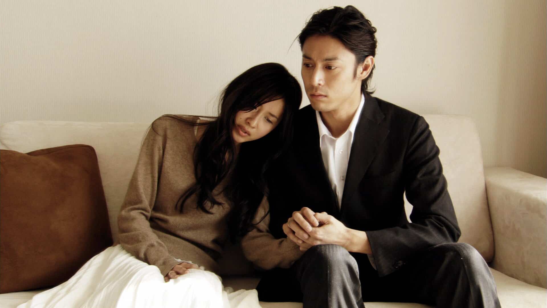 Kaho and Tomoya sit on a tan-colored couch in formal clothing, loosely clasping each other's hand. Kaho leans towards Tomoya, resting her head on his shoulder with a distraught expression. Tomoya stares pensively into off-screen space.